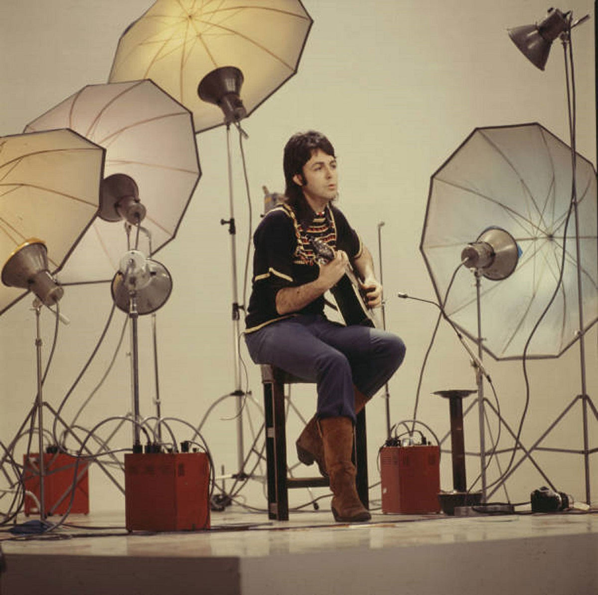 Paul McCartney seated with an acoustic guitar among photographic lights