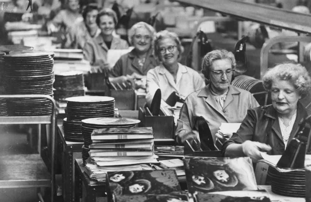 Black-and-white record production-line photograph with workers, stacks of discs and Rubber Soul sleeves