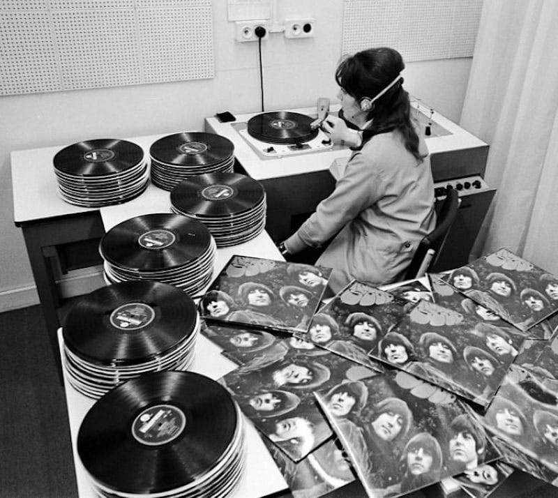 Black-and-white photograph of a headphone-wearing listener at record players beside stacks of discs and Rubber Soul sleeves